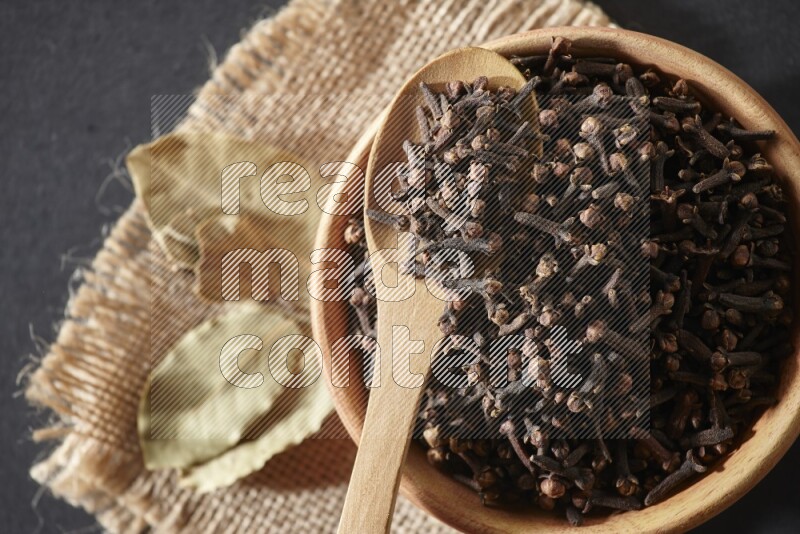 A wooden bowl, a wooden spoon full of cloves, and bay leaves (laurel) on a piece of burlap on a black flooring