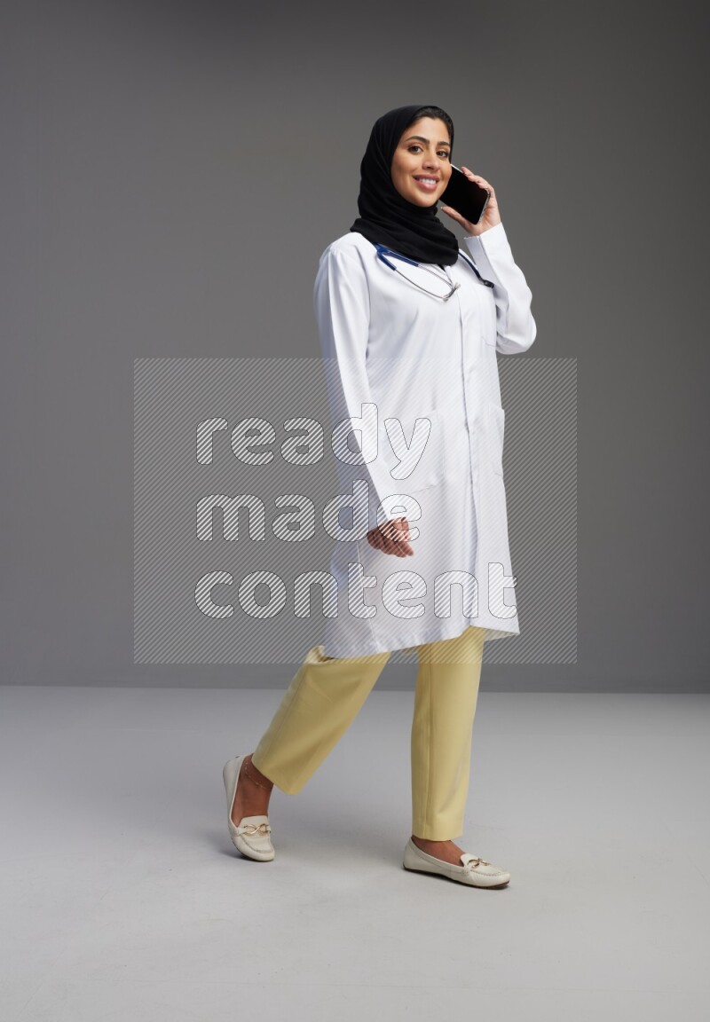 Saudi woman wearing lab coat with stethoscope standing talking on phone on Gray background
