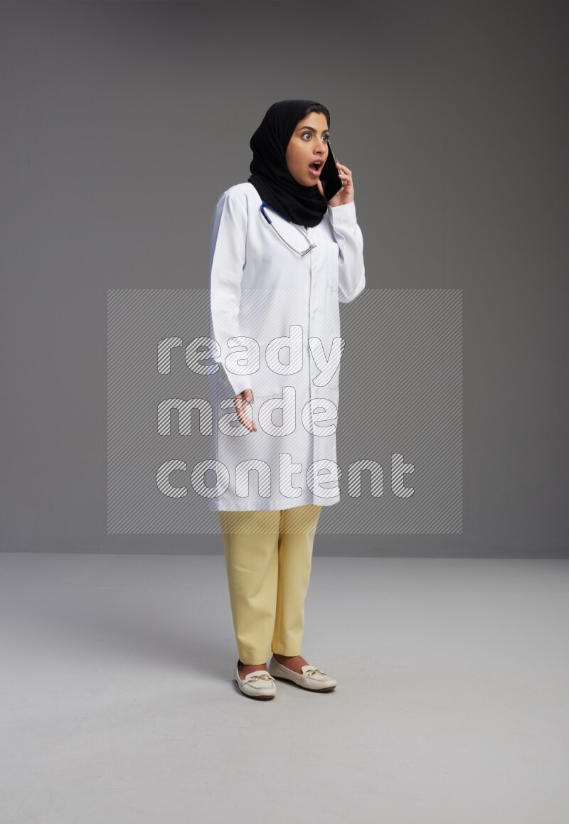 Saudi woman wearing lab coat with stethoscope standing talking on phone on Gray background