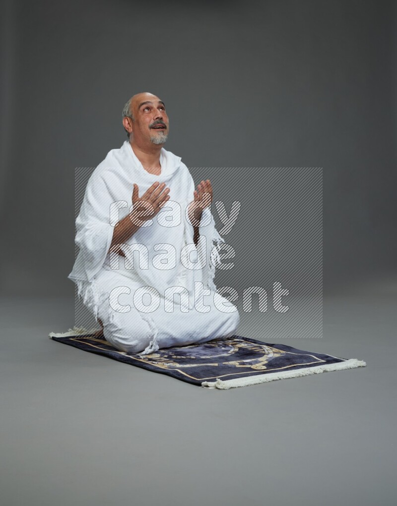 A man wearing Ehram sitting on prayer mat dua'a on gray background