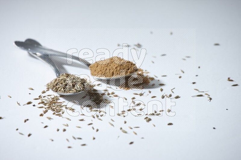 2 metal spoons full of cumin seeds and powder on white flooring